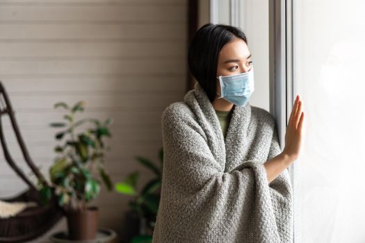 Sick asian girl in medical face mask standing by the window and yearning to go outside, being on quarantine, ill with covid-19 or flu.