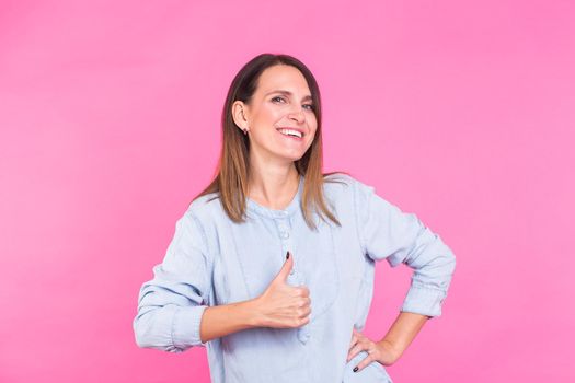 Portrait of a beautiful woman with long brown hair wearing blue cotton blouse, standing waist up smiling on a pink background.