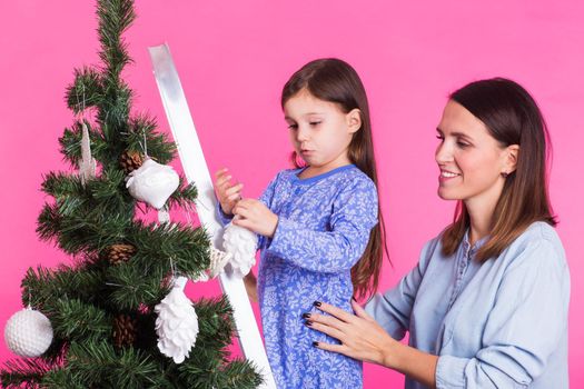 Holidays, family and christmas concept - mother and daughter decorating christmas tree on pink background