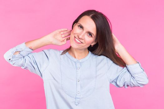 Portrait of a beautiful woman with long brown hair wearing blue cotton blouse, standing waist up smiling on a pink background.