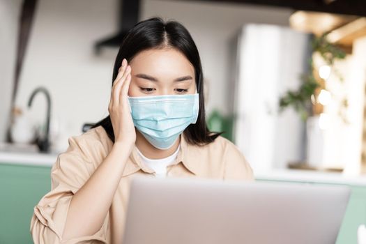 Young sick asian girl in medical face mask using laptop, working from home on quarantine.