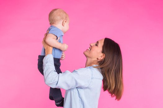 family, motherhood, parenting, people and child care concept - happy mother holds adorable baby over pink background.