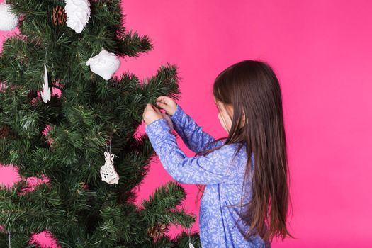 Children, holidays and christmas concept - little girl decorating christmas tree on pink background