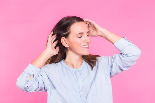 Portrait of a beautiful woman with long brown hair wearing blue cotton blouse, standing waist up smiling on a pink background.