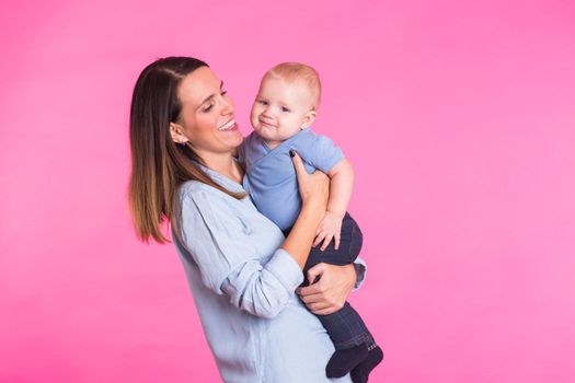 family, motherhood, parenting, people and child care concept - happy mother holds adorable baby over pink background.