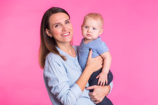 family, motherhood, parenting, people and child care concept - happy mother holds adorable baby over pink background.