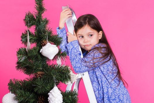 Children, holidays and christmas concept - little girl decorating christmas tree on pink background