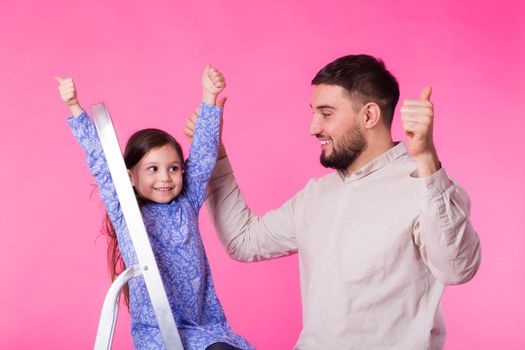 Father and her little daughter with thumbs up over pink background. Adult man and baby girl are happy.
