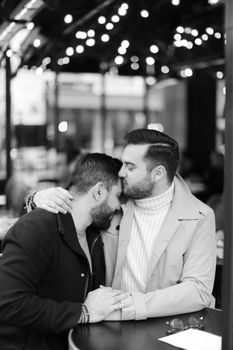 Black and white photo of caucasian gays sitting at street cafe, kissing and hugging. Concept of relationship and same sex couple.