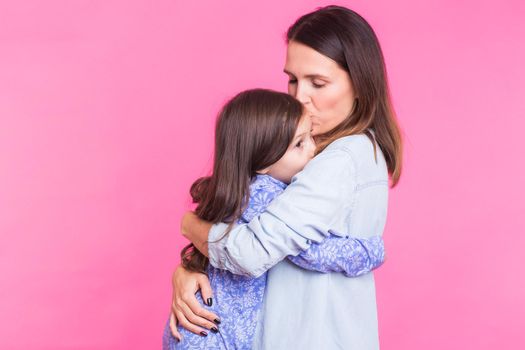 people, happiness, love, family and motherhood concept - happy little daughter hugging and kissing her mother over pink background.