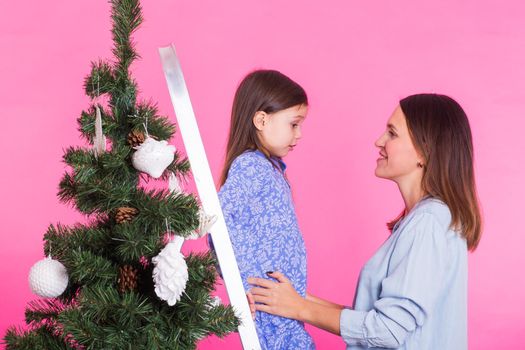 Holidays, family and christmas concept - mother and daughter decorating christmas tree on pink background