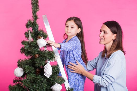 Holidays, family and christmas concept - mother and daughter decorating christmas tree on pink background