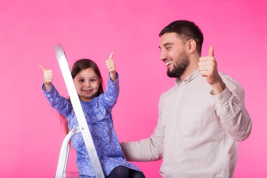 Father and her little daughter with thumbs up over pink background. Adult man and baby girl are happy.