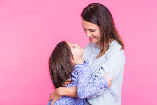 people, happiness, love, family and motherhood concept - happy little daughter hugging and kissing her mother over pink background.