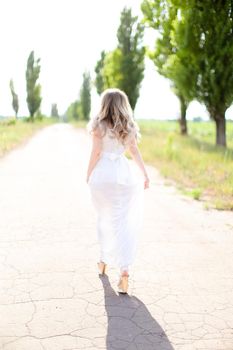 Back view of blonde caucasian bride wearing white dress walking on road. Concept of wedding photo session and fiancee.