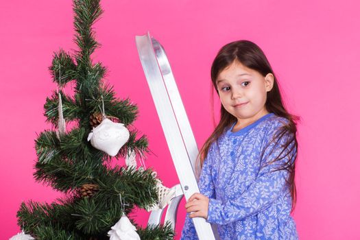 Children, holidays and christmas concept - little girl decorating christmas tree on pink background