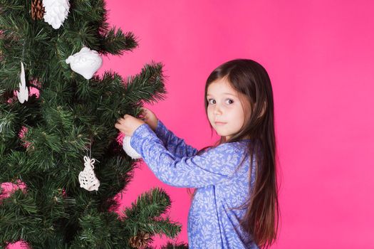 Children, holidays and christmas concept - little girl decorating christmas tree on pink background