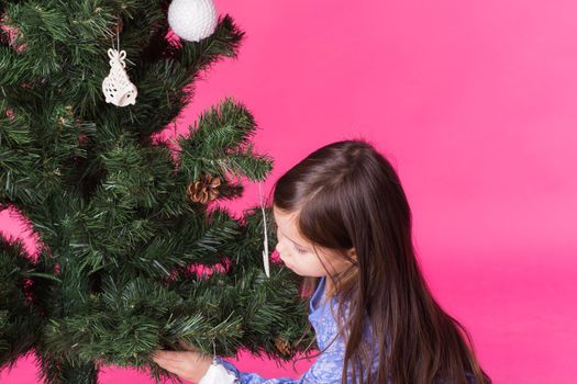 Children, holidays and christmas concept - little girl decorating christmas tree on pink background