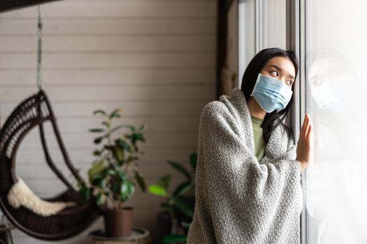 Sad asian girl in medical face mask looking outside window and yearning go for walk, being ill, catching covid 19 or flu.