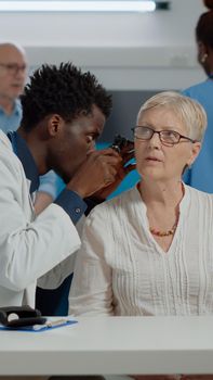 Healthcare specialist holding professional otoscope for ear examination of elderly patient at desk in doctors office. Otologist using tool on caucasian senior woman with infection at clinic