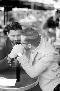 Black and white photo of two young handsome gays talking at street cafe and holding hands. Concept of same sex couple and lgbt.