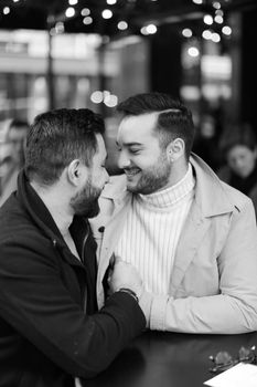 Black and white photo of caucasian gays sitting at street cafe, smiling and hugging. Concept of relationship and same sex couple.