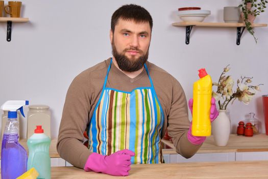 Image of a young Caucasian bearded man in the kitchen, he is preparing to do the cleaning, showing his favorite detergent