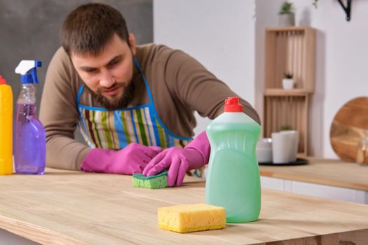 Image of a young Caucasian bearded man in the kitchen, he is desperately trying to clean a table, using detergent