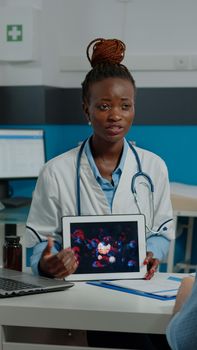 Doctor of african american ethnicity with virus animation on digital tablet showing possible disease from covid 19 in medical cabinet. Old patient looking at coronavirus bacteria