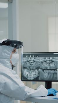 Professional dentistry staff with ppe suit looking at teeth x ray in dental cabinet. Assistant examining teeth radiography on computer monitor while doctor preparing for consultation