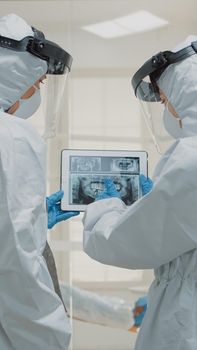 Professional orthodontists looking at dental x ray on digital tablet at clinic wearing protection suits during coronavirus epidemic. Dentists examining teeth radiography on scan device