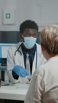 Adult medic discussing medical treatment with senior patient sitting at desk with glass protection against coronavirus. Doctor and old woman with face masks at appointment for annual checkup