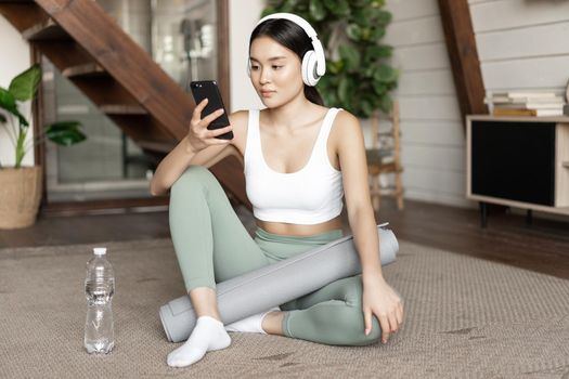 Young asian woman with water bottle, listening music or podcast in headphones during fitness at home, sitting on floor before exercising.