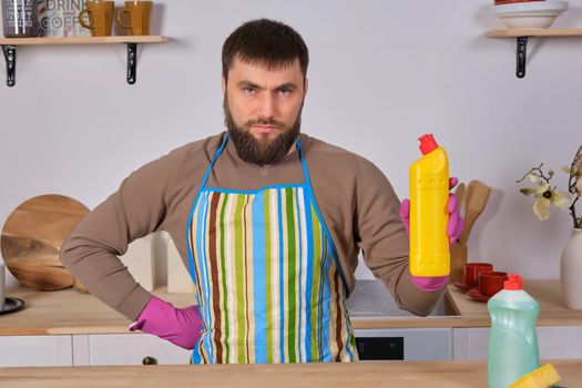 Housecleaning concept. Young bearded man in kitchen makes some advertising. He promotes detergent, holding it in one hand and shows at camera