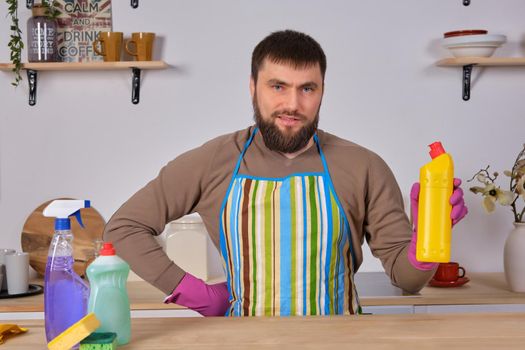Housecleaning concept. Young bearded man in kitchen makes some advertising. He promotes detergent, holding it in one hand and shows at camera