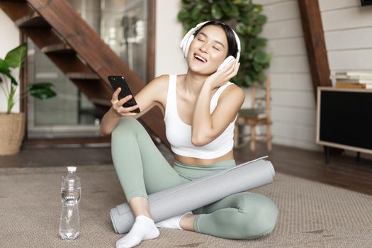 Happy asian girl in fitness clothing sitting at home on floor, listening music and workout, holding smartphone and singing during training session.