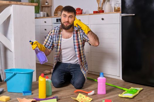 Housecleaning concept. Caucasian handsome young bearded man in checkered shirt sits on the floor of his kitchen and tries to clean it using all of his detergents, rags, washcloths and brushes