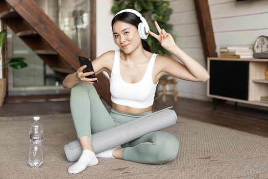 Happy sporty girl sitting on floor at home and taking selfie during workout, wearing headphones and holding smartphone. Concept of sport and lifestyle