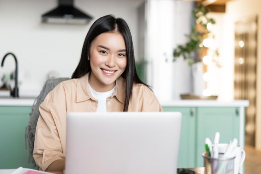 Young asian girl freelancer working remotely from home. Woman with laptop studying indoors from her kitchen, smiling at camera.