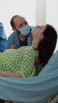 Obstetrician and african american nurse delivering baby in hospital ward assisting pregnant patient with painful labor. Young husband holding hand helping screaming wife with pushing