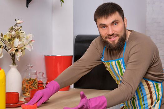 Image of a young Caucasian bearded man in the kitchen, he is desperately trying to clean a table, using detergent