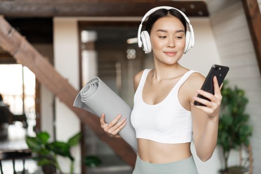 Young asian woman standing at home in headphones, listening music and going workout on floor mat, practice yoga or meditate in living room.