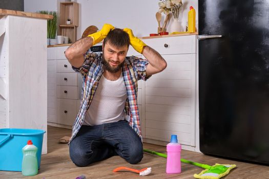 Housecleaning concept. Caucasian handsome young bearded man in checkered shirt sits on the floor of his kitchen and tries to clean it using all of his detergents, rags, washcloths and brushes