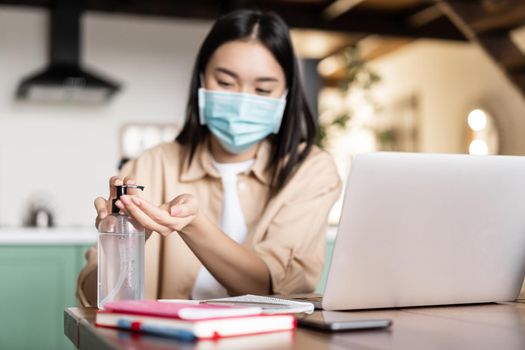 Young asian girl student using hand sanitizer in medical face mask, working on laptop computer remotely. Freelancer works at coworking space.