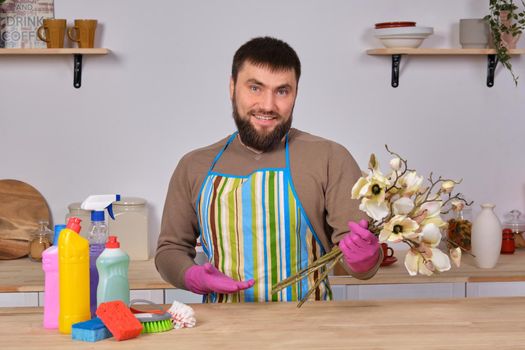 Image of a young Caucasian bearded man in the kitchen, he makes little presentation of all his detergents, preparing for big cleaning. Man caring for flowers