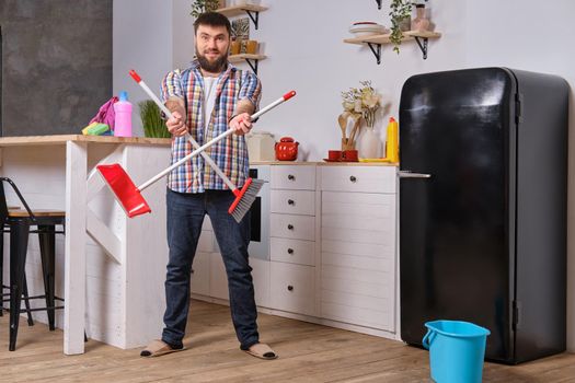 Professional photo of Young handsome bearded man in the kitchen, wearing checkered shirt. He wants to sweep the floor but has some difficulties with a dustpan and a broom.