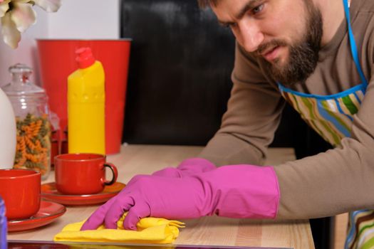 Close up Image of a young Caucasian bearded man in the kitchen wearing apron. He tries to clean the cooking surface and near. Different detergents used