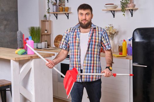 Professional photo of Young handsome bearded man in the kitchen, wearing checkered shirt. He wants to sweep the floor but has some difficulties with a dustpan and a broom.
