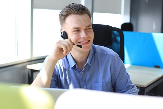 Cheerful young support phone male operator in headset, at workplace while using laptop, help service and client consulting call center concept.