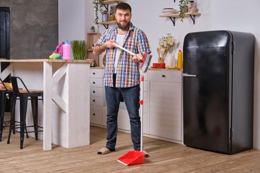 Professional photo of Young handsome bearded man in the kitchen, wearing checkered shirt. He wants to sweep the floor but has some difficulties with a dustpan and a broom.